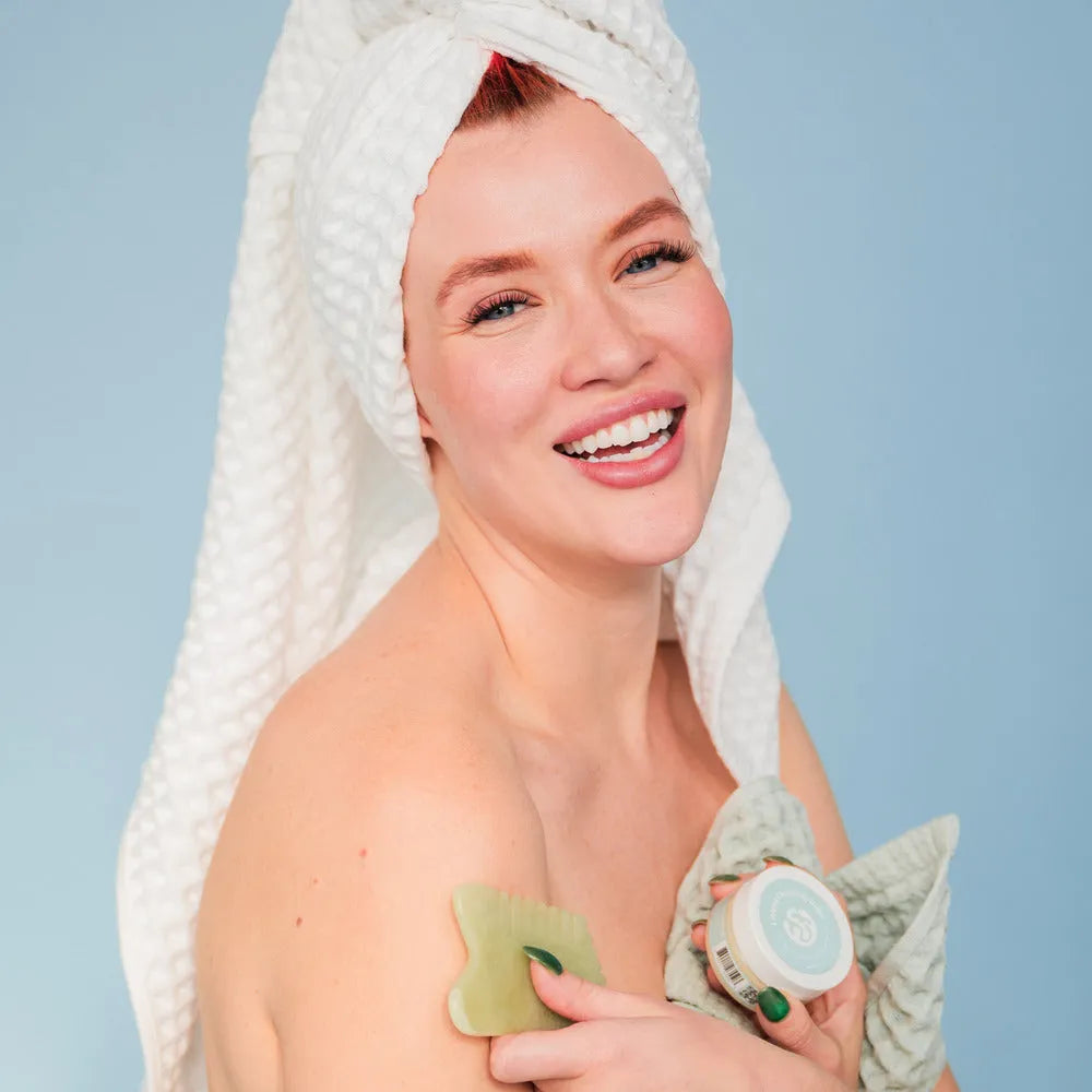 A radiant woman wrapped in a white towel smiles while holding a jade gua sha tool and skincare product, highlighting a joyful self-care routine.