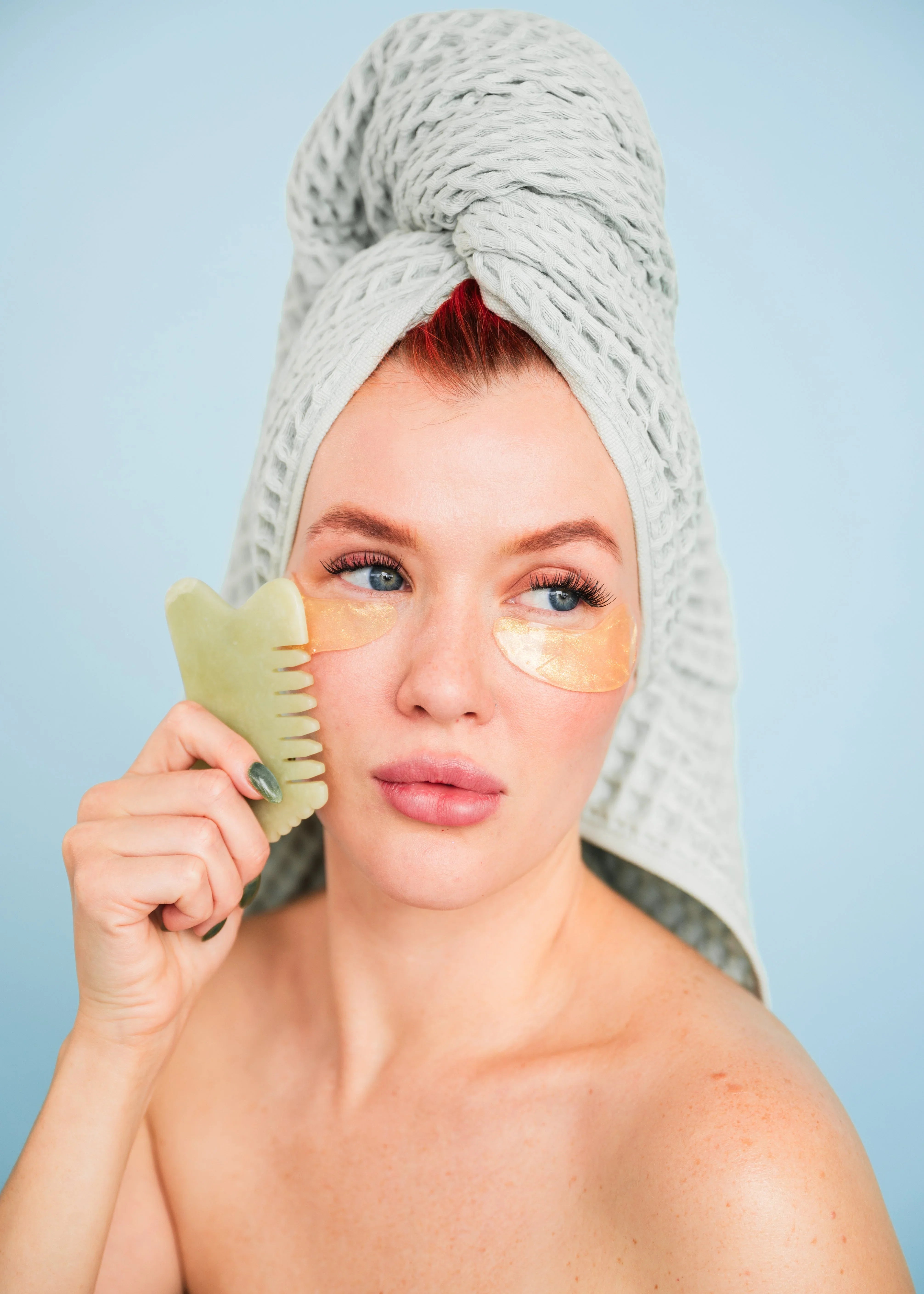 Woman with towel on head using jade gua sha tool and under-eye patches, Bohemia Skin spa care