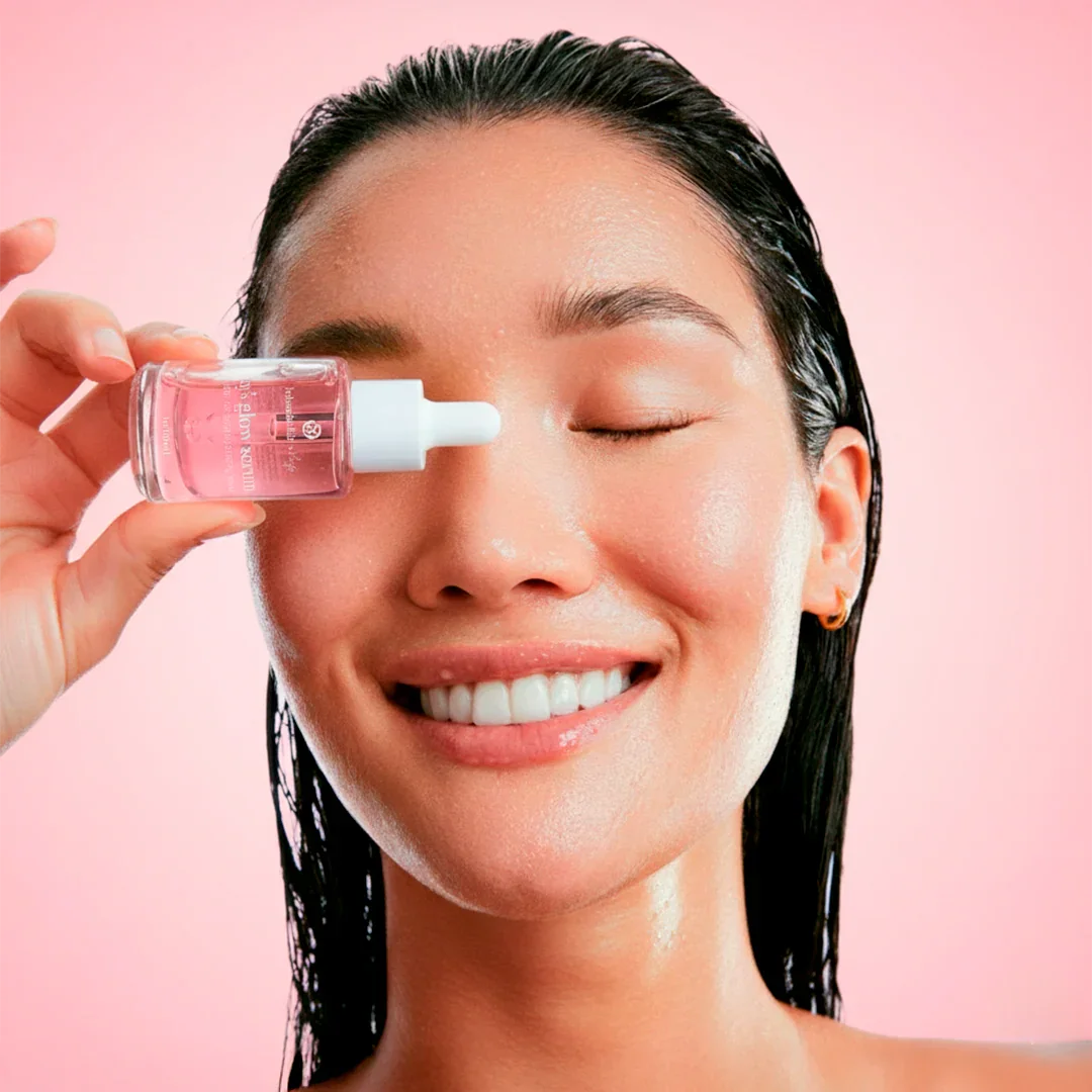 Smiling woman with glowing skin holding Bohemia Skin serum bottle against pink background