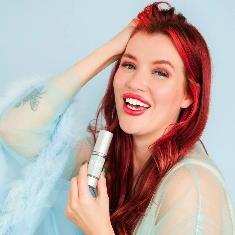 A smiling woman with red hair holds Bohemia's YTH Serum, showing off radiant, glowing skin in a sheer blue outfit against a soft blue background.